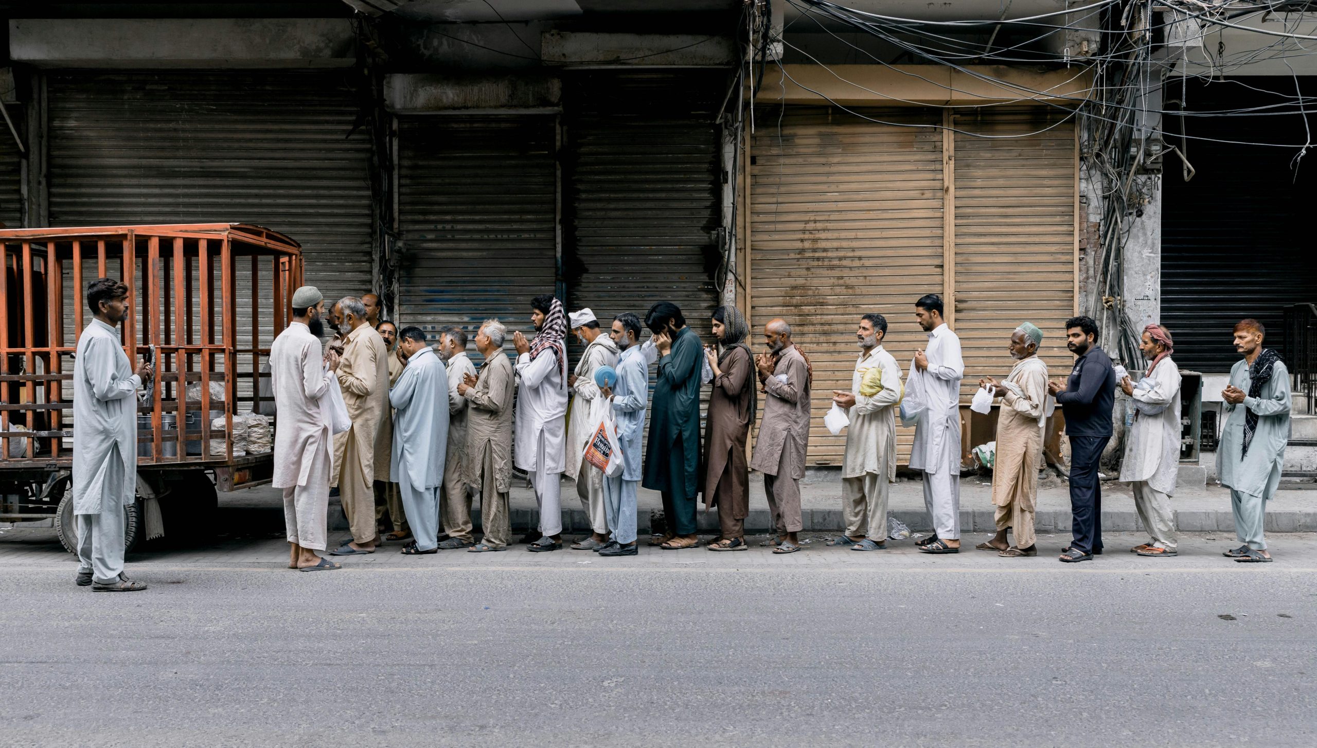 A group of adults standing in line for charity aid on a city street in Faisalabad, Pakistan.